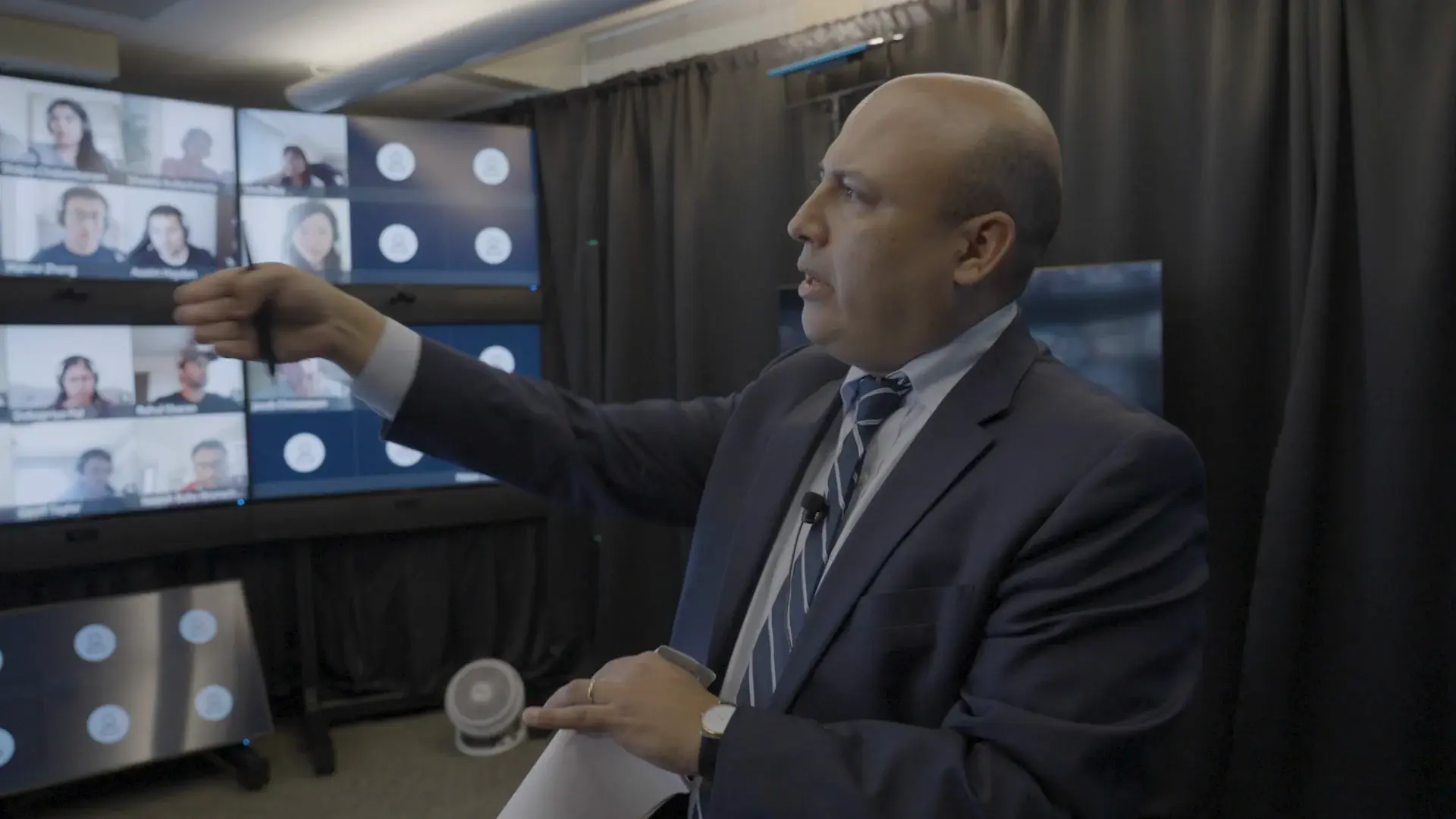 A man points at a collection of screens showing students on a Zoom meeting